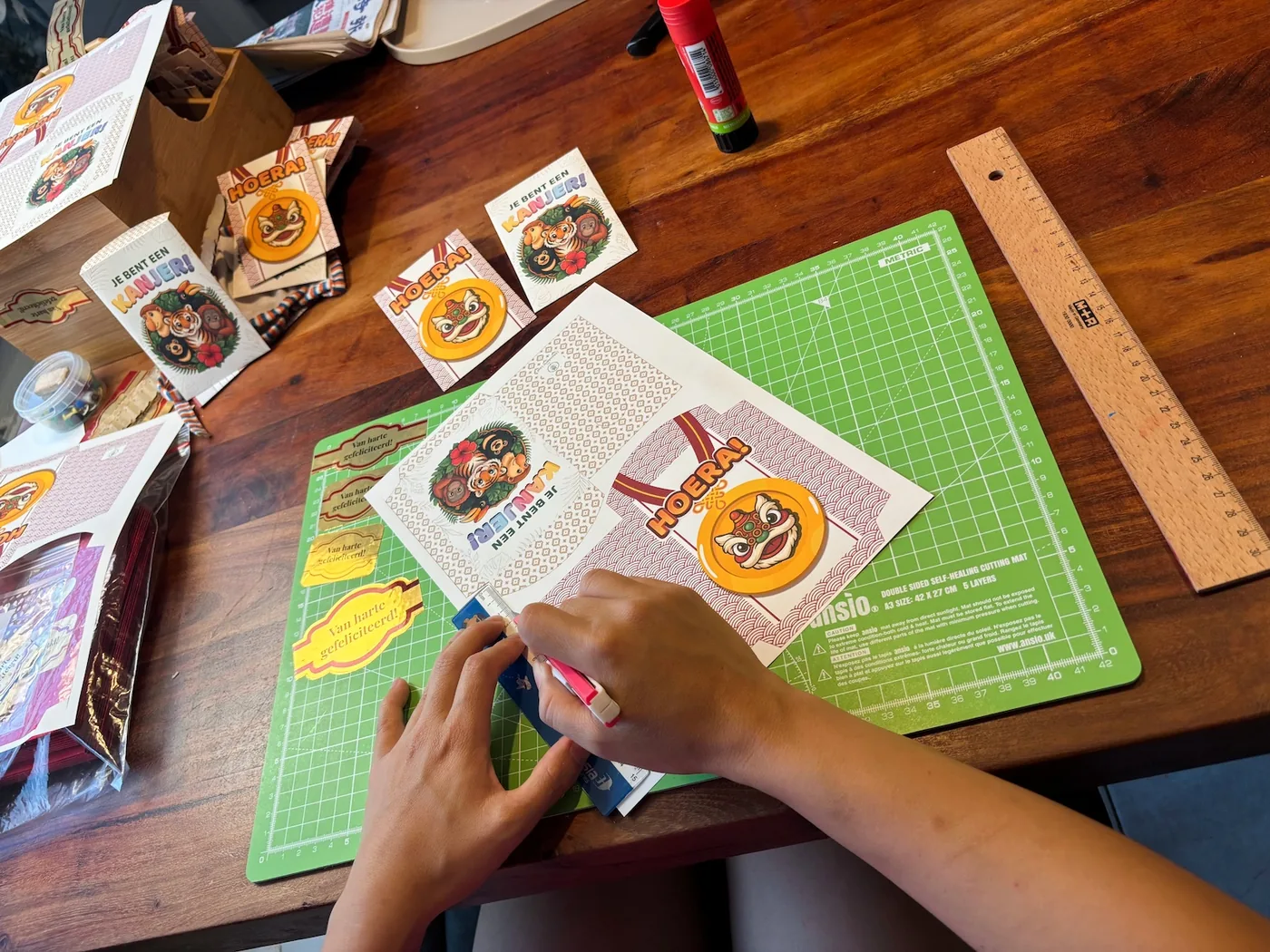 Cutting and folding angpao by hand at the kitchen table, surrounded by paper and designs